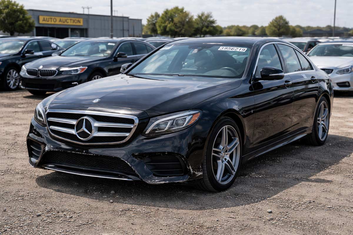 lack Mercedes-Benz E-Class with slight front bumper damage, polished and parked in an active salvage auction yard, with auction lot numbers and urban backdrop.